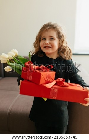 Close up incredible little girl with wavy hairstyle is holding big presents and flowers. Charming little cute girl is posing over isolated background. High quality photo