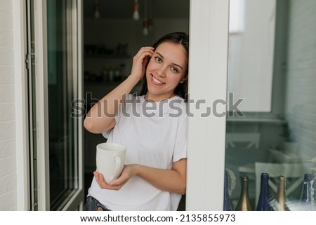 Adorable lovely woman smiling and touching her hair while posing at camera with morning cup with coffee at home in the kitchen. High quality photo