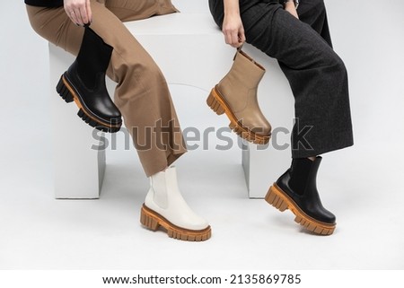Two girls in a coat and tights posing with their feet in boots in the studio on a white background, holding dark leather boots boots in their hands Royalty-Free Stock Photo #2135869785