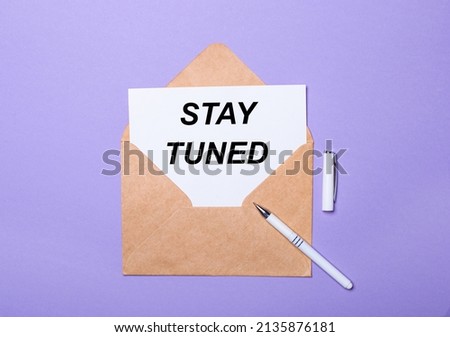 On a purple background, a white pen, a craft envelope and a white card with the text STAY TUNED. View from above.