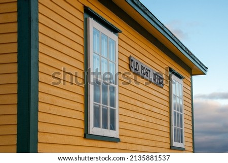 A wooden sign with black letters spelling old post office is affixed to the exterior of a yellow building with green trim. There are vintage glass windows with white trim on the corner of the building
