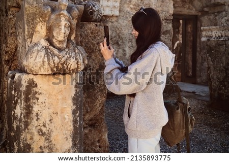 Young caucasian woman taking photo of old ruined ancient stone sculpture on smartphone. Selective focus.