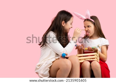Picture of two sisters preparing for the Easter party wearing bunny ears and painting the nose, making fun together.