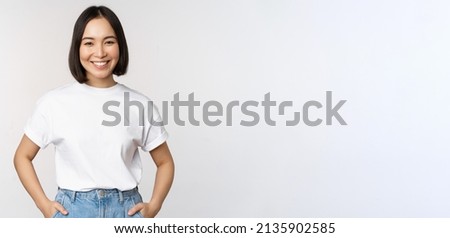 Lifestyle. Happy modern asian girl, smiling and looking happy at camera, posing in white tshirt and jeans, studio background