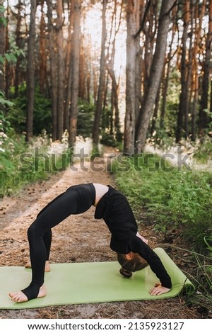 A diligent, beautiful girl of preschool age, a professional child in a black uniform is engaged in gymnastics, yoga on a green rug in the forest in nature, standing on the bridge.