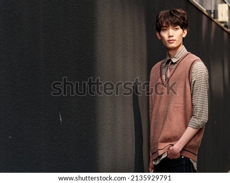 Portrait of handsome Chinese young man with curly black hair in plaid shirt and wool vest standing against black wall background in sunny day, male fashion, cool Asian young man lifestyle.
