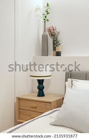Fabric upholstered bedroom headboard attached to plasterboard construction with vases of flowers and light wood nightstand with blue lamp