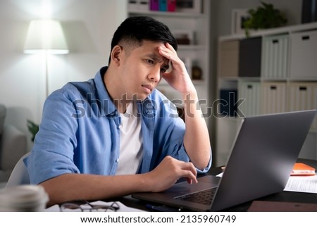 Unhappy young Asian man, entrepreneur, student, tutor looking at laptop screen with serious and worried face and with hand on forehead when working or learning from home. Royalty-Free Stock Photo #2135960749