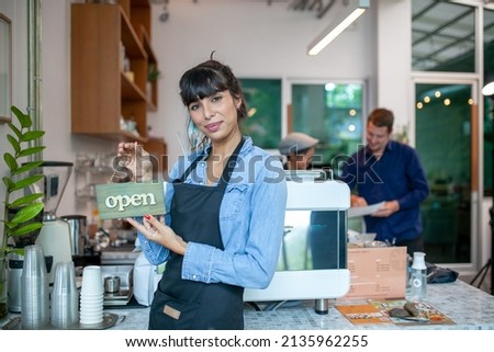 Portrait of smiling owner standing at her cafe with open signboard. Coffee owner standing in front of coffee shop to welcome customer and open the coffee shop in morning.
