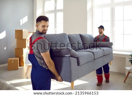 Two professional relocation service workers in overalls move sofa in customer's apartment. Movers carry sofa, cardboard boxes and assembling furniture. Moving and delivery company services. Royalty-Free Stock Photo #2135966153