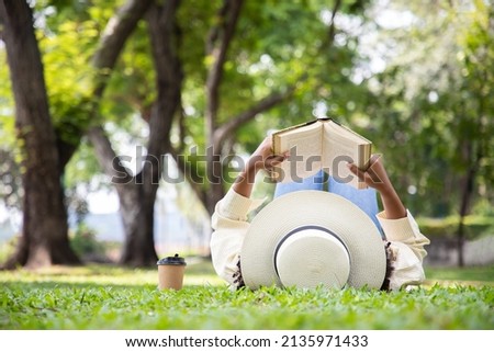 Woman reading book in grass under tree with cup of coffee. Relax in summer time holiday laying on the grass field comfortable feeling. Royalty-Free Stock Photo #2135971433