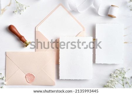 Blank white invitation cards with pink envelopes, gypsophila branches, wax seal stamp, silk ribbon. Wedding stationery set. Flat lay, top view, copy space.