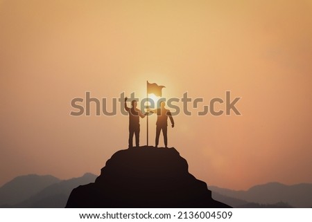 Business Success, Leadership and Success and Goal Concepts. Silhouette of businessman with flags on mountain peaks above sky and sunset background. Royalty-Free Stock Photo #2136004509