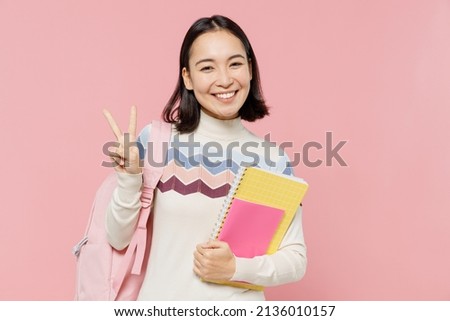Friendly fun teen student girl of Asian ethnicity wear sweater backpack hold books showing victory sign isolated on pastel plain light pink color background. Education in university college concept
