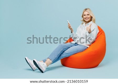 Full body fun elderly woman 50s wearing casual striped shirt sit in bag chair use hold mobile cell phone show thumb up isolated on plain pastel light blue color background. People lifestyle concept