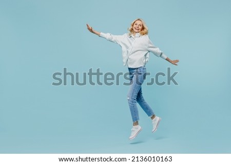 Full body elderly woman 50s wearing casual striped shirt looking camera jump hilg with outstretched hands like fly run isolated on plain pastel light blue color background. People lifestyle concept.