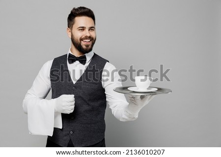 Young fun barista male waiter butler man wear white shirt vest elegant uniform work at cafe hold give plate coffee cup isolated on plain grey background studio portrait. Restaurant employee concept Royalty-Free Stock Photo #2136010207