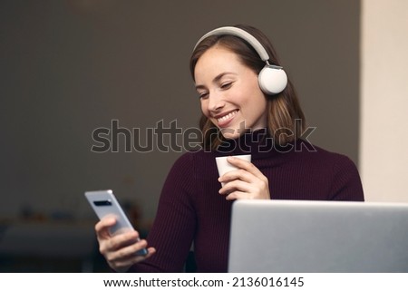 Happy and smiling brunette girl with headphones sitting in front of laptop with a cup of coffee, while looking at her smartphone, listening to music or podcasts	