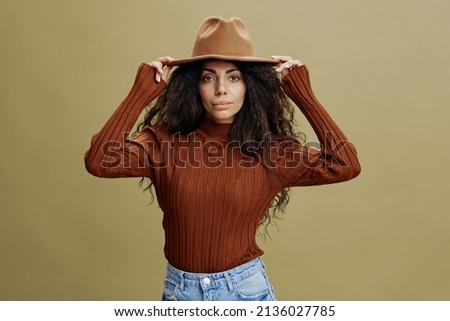 Serious confident stylish curly Latin female in brown hat, looking at camera, holding hands on hat, isolated green background. Mockup. Copy space clothing fashion brands, free place for your ad. 