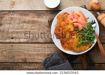 Potato pancakes. Fried homemade potato pancakes or latkes with cream, green onions, microgreens, red salmon and sauce in rustic plate on old wooden table background. Rustic style. Top view.