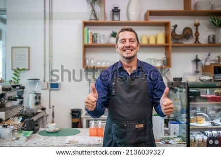 Portrait of smiling owner man standing at his cafe with thumbs up hand. Coffee owner standing with apron in coffee shop to welcome customer.