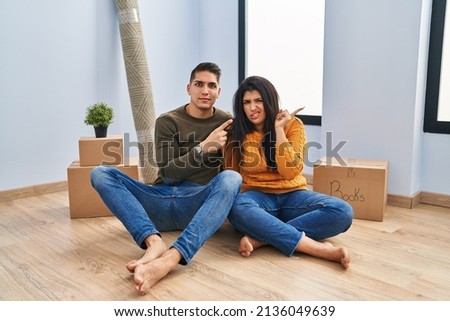 Young couple sitting on the floor at new home pointing aside worried and nervous with forefinger, concerned and surprised expression 