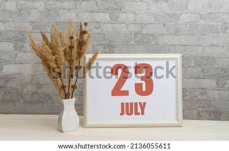 july 23. 23th day of month, calendar date. White vase with ikebana and photo frame with numbers on desktop, opposite brick wall. Concept of day of year, time planner, summer month.
