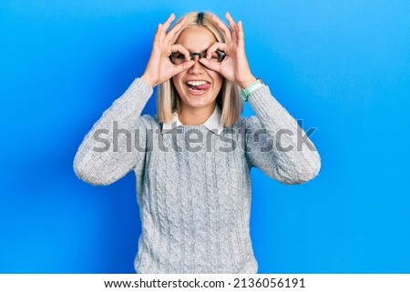 Beautiful blonde woman wearing glasses doing ok gesture like binoculars sticking tongue out, eyes looking through fingers. crazy expression. 