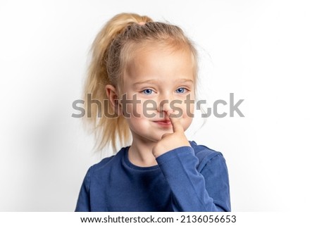 A little girl with blue eyes cheerfully touches her nose with her finger. Royalty-Free Stock Photo #2136056653