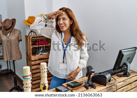 Middle age hispanic woman working as manager at retail boutique very happy and smiling looking far away with hand over head. searching concept.  Royalty-Free Stock Photo #2136063063