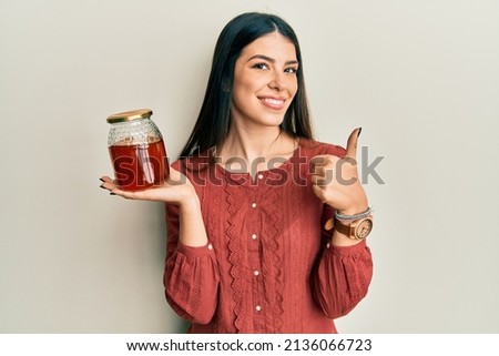 Young hispanic woman holding jar with honey smiling happy and positive, thumb up doing excellent and approval sign 