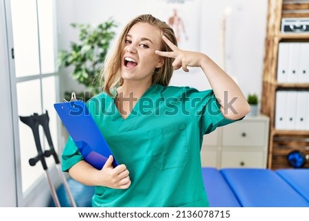 Young caucasian woman working at pain recovery clinic doing peace symbol with fingers over face, smiling cheerful showing victory 