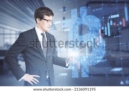 Attractive young caucasian businessman holding tablet with creative glowing bitcoin grid forex chart hologram on blurry office interior background. Cryptocurrency and growth concept. Double exposure