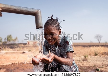 Happy little African girl crouching in front of a waterhole in an arid environment somewhere in the subsaharn region, drinking lots of fresh water from her cupped palms; world water day icon Royalty-Free Stock Photo #2136097877
