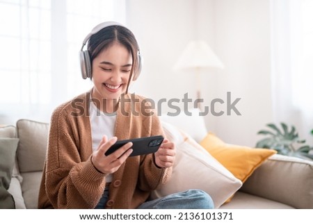 Happy young asian woman relaxing and listening music and watches movie on mobile phone with headphones while sitting on a couch in the morning at home Royalty-Free Stock Photo #2136101371