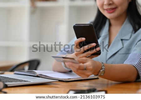 Cropped view of a pretty smiling Asian woman sitting in the office holding a credit card and a smart phone, for finance and technology concept.
