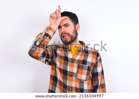 Funny young caucasian man wearing plaid shirt over white background makes loser gesture mocking at someone sticks out tongue making grimace face.