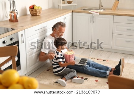 Little boy and his grandfather with tasty popcorn using laptop in kitchen