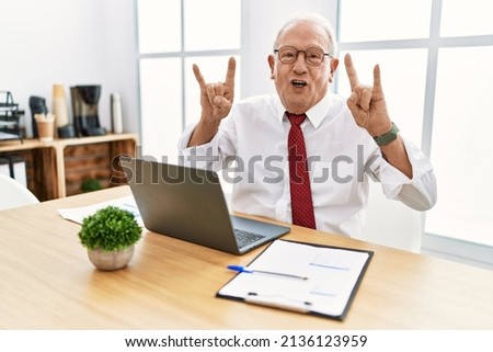 Senior man working at the office using computer laptop shouting with crazy expression doing rock symbol with hands up. music star. heavy music concept. 