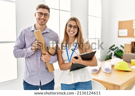 Two young architect smiling happy holding blueprints and clipboard at architecture studio.
