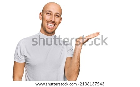 Bald man with beard wearing casual white t shirt smiling cheerful presenting and pointing with palm of hand looking at the camera. 