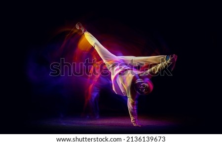 Solo dance. Young flexible sportive man dancing hip-hop in white outfit on dark background in mixed yellow neon light. Beauty, sport, youth, action, moves. Dancer shows breakdance figures Royalty-Free Stock Photo #2136139673