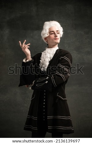 Young man in image of Amadeus Mozart, medieval person isolated on dark vintage background. Retro style, comparison of eras concept. Elegant male model as historical character, great music compose Royalty-Free Stock Photo #2136139697