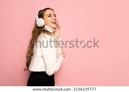 Happy, positive. Young beautiful smiling girl in warm white sweater listeninf to music in headphones isolated on pink background. Concept of emotions, facial expression, youth, aspiration, sales.