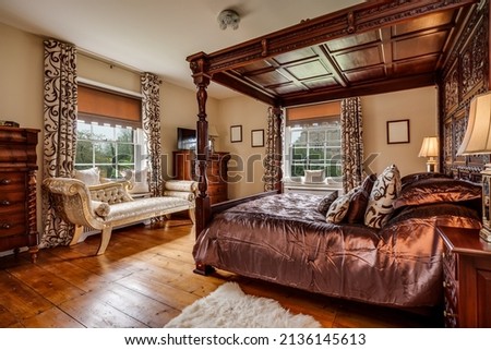 Furnished bedroom within former victorian rectory with ornate carved four poster bed and matching period sideboard drawers, chaise longe, side tables with lamps and exposed timber floor Royalty-Free Stock Photo #2136145613