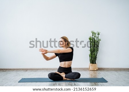 A woman trains sitting on a yoga mat, stretching her arm to the side, stretching muscles during training, preparing the body for exercises. High quality photo Royalty-Free Stock Photo #2136152659