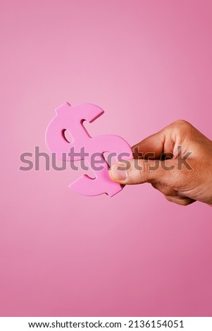 closeup of the hand of a man grabbing a pink dollar sign on a pink background, depicting the pink money concept or pink capitalism