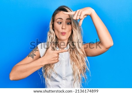 Beautiful young blonde woman doing picture frame gesture with hands making fish face with mouth and squinting eyes, crazy and comical. 