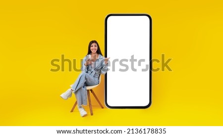 Check This. Excited lady sitting on chair using cell pointing finger at big giant smart phone with empty white screen, presenting new cool app, free copy space mock up, website design banner