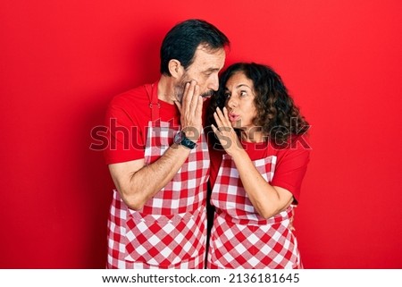 Middle age couple of hispanic woman and man wearing cook apron hand on mouth telling secret rumor, whispering malicious talk conversation 